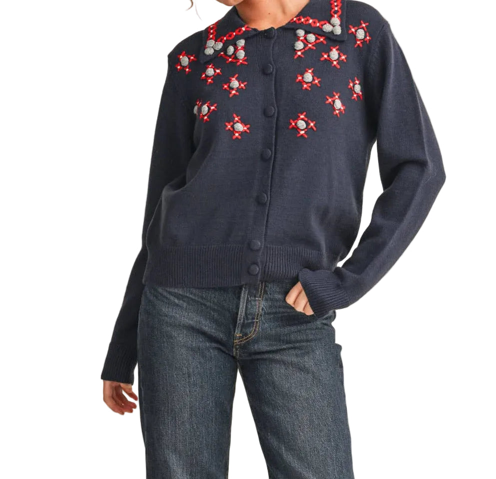 A woman modeling a navy blue button-up shirt with red and white floral embroidery on the chest, paired with dark blue jeans and red flats, against a solid beige background.