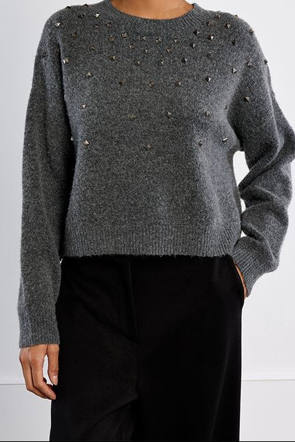 A woman with curly dark hair wearing a gray knit sweater with small metallic embellishments across the chest and black trousers, standing against a plain light gray studio background.