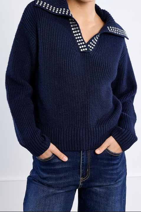 A woman wearing a navy blue ribbed knit sweater with a studded collar detail, paired with dark blue jeans, against a plain white studio background.