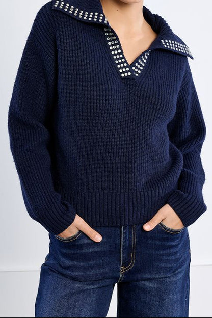 A woman wearing a navy blue ribbed knit sweater with a studded collar detail, paired with dark blue jeans, against a plain white studio background.