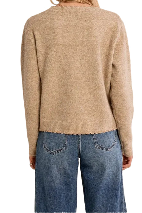 A woman viewed from behind wearing a beige knit sweater with a scalloped edge and blue jeans, set against a plain white studio background.