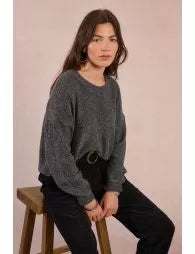 Woman sitting on a wooden stool wearing a gray sweater and black pants against a plain background