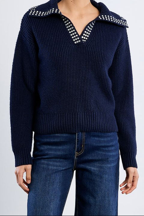 A woman modeling a navy blue ribbed knit sweater with a wide collar and silver rhinestone trim, paired with dark blue jeans against a light gray studio background.