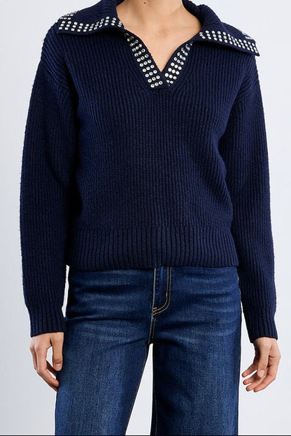 A woman modeling a navy blue ribbed knit sweater with a wide collar and silver rhinestone trim, paired with dark blue jeans against a light gray studio background.