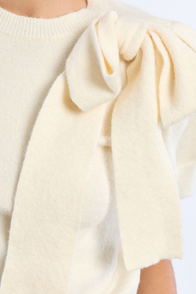 Close-up of a cream-colored knit sweater featuring a large decorative bow detail on the shoulder.