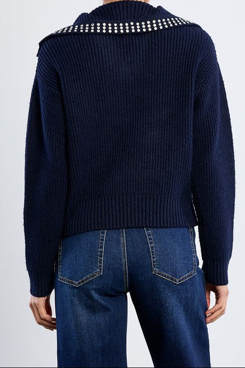 A woman viewed from the back wearing a navy ribbed knit sweater with a row of white rhinestone embellishments across the shoulders, paired with blue jeans, against a light gray studio background.