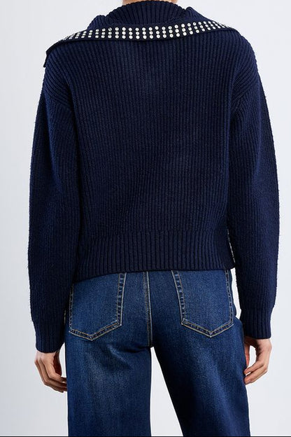 A woman viewed from the back wearing a navy ribbed knit sweater with a row of white rhinestone embellishments across the shoulders, paired with blue jeans, against a light gray studio background.